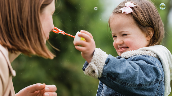 A cheerful child with Down syndrome sitting with an adult, blowing bubbles outdoors. This image illustrates the enhanced member supports offered by Highmark Healthy Kids.
