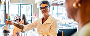 A middle aged person smiles as they sit at a counter in a restaurant while speaking with someone else.