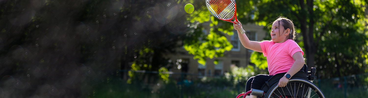 A young girl in a wheelchair smiling as she plays tennis outdoors, reaching up with a racket to hit a tennis ball.