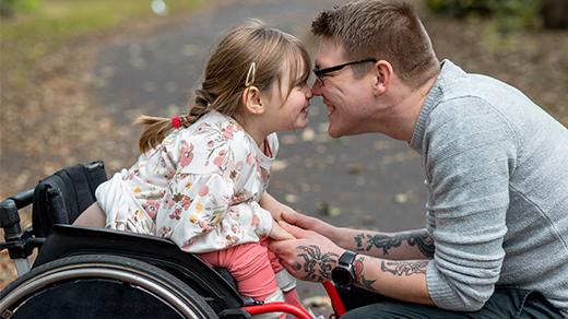 A young girl in a wheelchair smiling joyfully and touching noses with a friend kneeling in front of her.