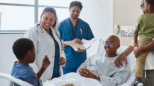 A smiling doctor and a nurse speaking with a patient resting in a hospital bed, surrounded by family members.