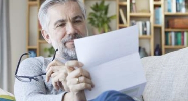 Man with grey hair and beard holding glasses sits on his couch at home reviewing Highmark Medicare Plans letter