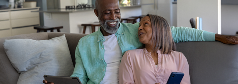 An older black couple on a couch, smiling and engaged with their smart devices, against a modern kitchen backdrop, illustrates "How Medicare Advantage Compares to Other Medicare Add-Ons."