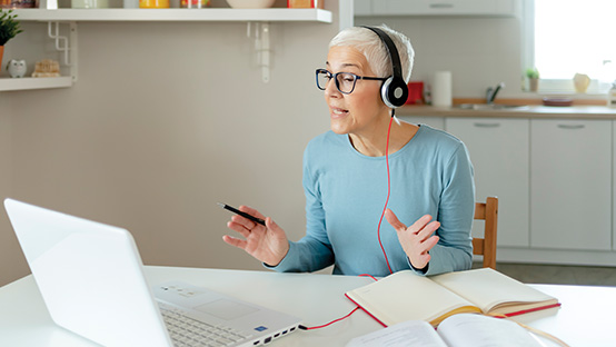 A senior woman wears headphones and is talking and looking at an open laptop; two books are open in front of her