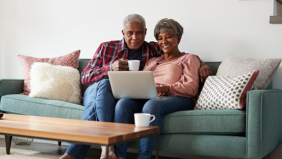 A senior man and senior woman sit on a couch and look at the laptop that is sitting open on the woman’s lap.