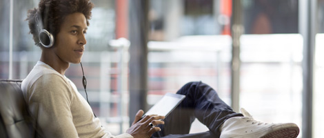 A young Black man sits relaxed, listening to music on headphones while looking at a tablet.