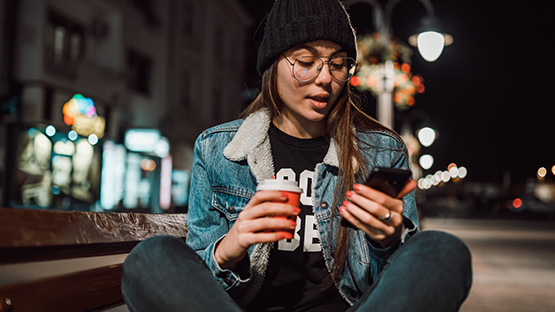 A young female adult on the street at night with a coffee and a mobile device to illustrate the many available crisis hotlines.