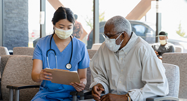 female medical person speaking with elderly man sitting in waiting room wearing masks