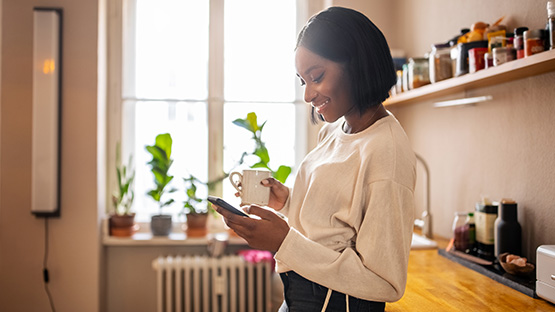 woman standing in kitchen holding a cup of coffee and reading her smart phone