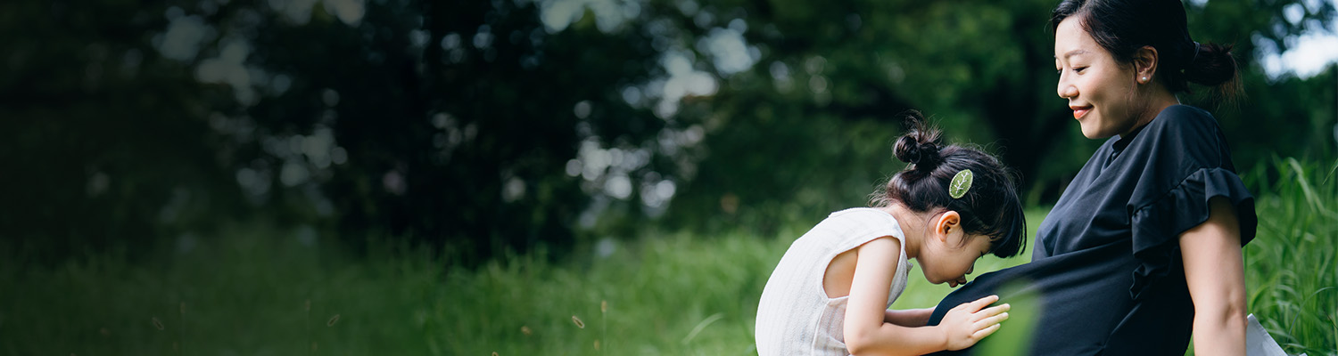 Pregnant mother sitting in grass with toddler daughter sitting on lap kissing pregnant belly