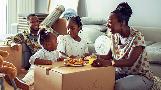 Mother and father with two young daughter moving into a new home using a moving box as table to have lunch