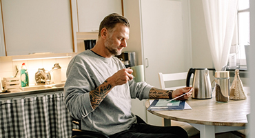 Older man sitting at table in kitchen drinking coffee and looking at bills