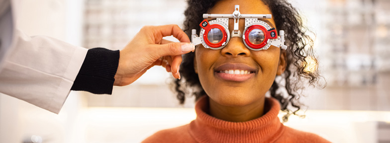 A young adult smiles while a medical professional in a black and white long-sleeved shirt helps them with an eye exam machine.