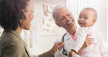 Woman doctor holding a baby, the baby's mother is next to them and they are all smiling
