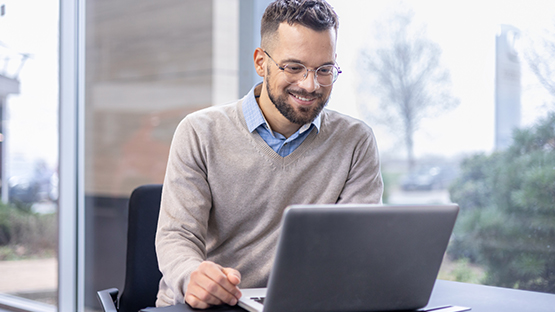 A young man is in an office, wearing casual work attire and looking at his laptop. 