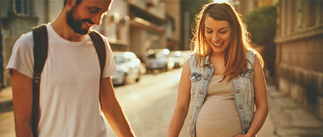 A young couple walk down a city street, holding hands. The woman is pregnant. 