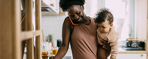 Woman in kitchen, on the phone and holding her baby. 