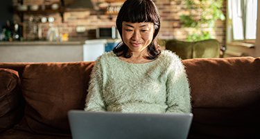 A woman is sitting on her sofa in her living room with a laptop on her lap. She is looking at the screen and has a natural smile on her face.
