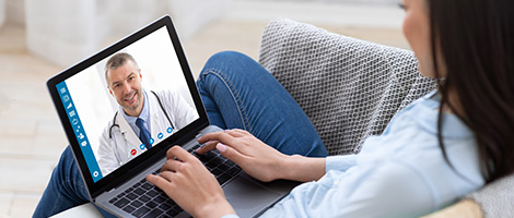 Over-the-shoulder view of a woman sitting on a sofa, at home, with a laptop on her lap. The laptop screen shows that she is in a virtual doctor visit. 