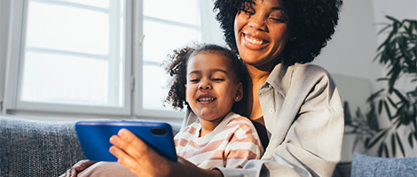 A woman sitting on the living room sofa with her daughter on her lap. They are both looking at a phone, talking to someone via video chat. 