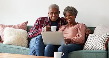 A senior man and senior woman sit on a couch and look at the laptop that is sitting open on the woman’s lap