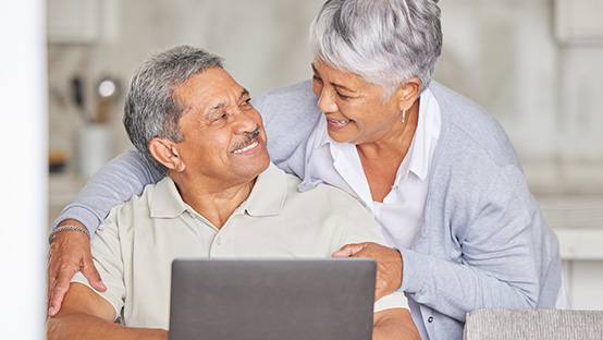 A standing senior woman leans over a seated senior man and the two people are smiling at each other