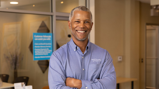 A smiling Black man stands with arms folded, ready to chat about Medicare